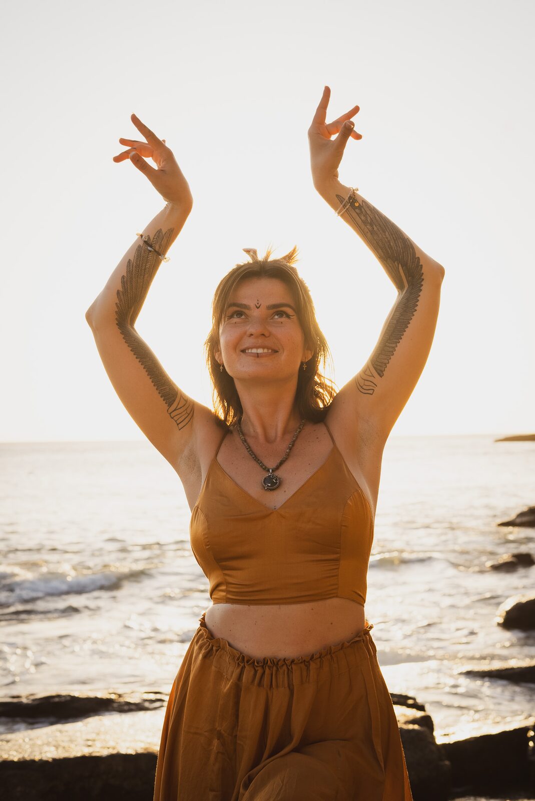 TSA Revna smiling with both arms raised above her head against a shimmering ocean-sunset horizon, eagle-wing tattoos visible, ochre golden-hour light