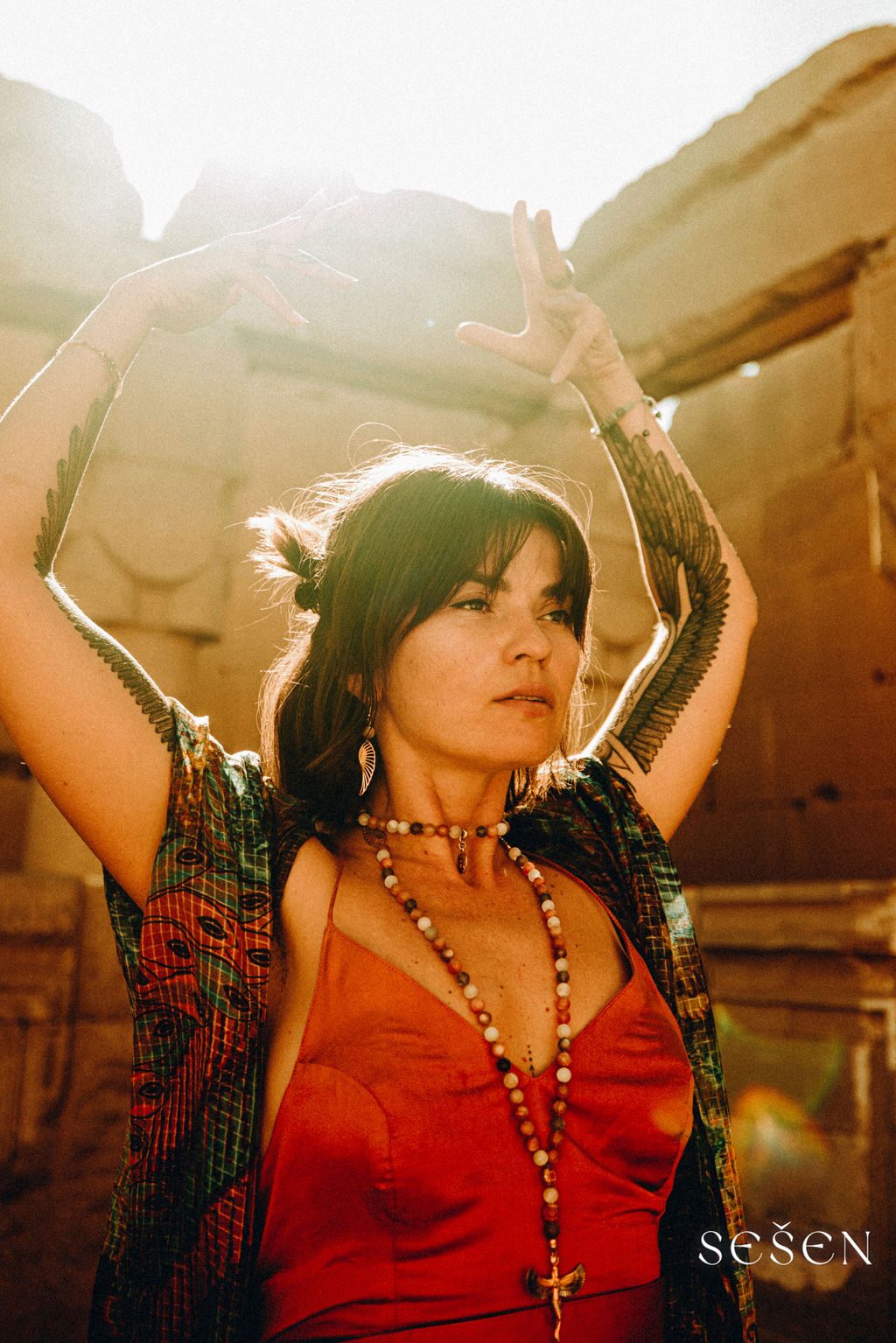 TSA Revna in a red silk slip dress with arms raised overhead among ancient stone ruins, eagle-wing tattoos along her arms, warm desert sunlight