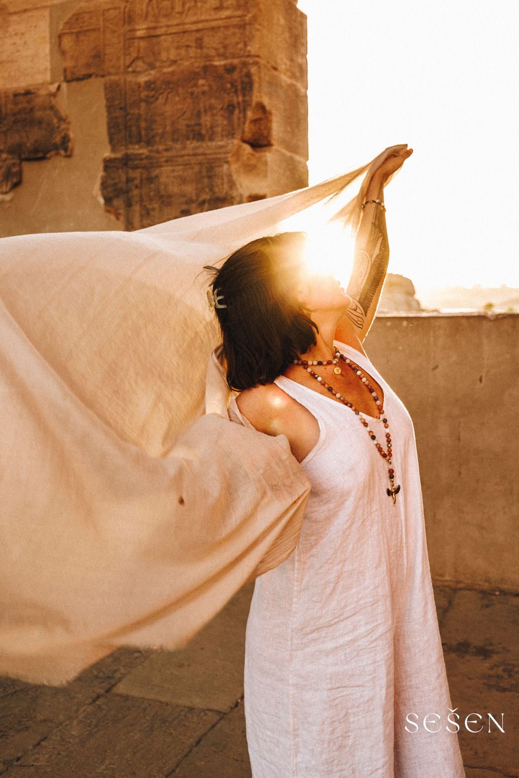 TSA Revna lifting a flowing sheer veil above her head at an ancient Egyptian temple, golden sun flaring through stone columns, mala beads, hieroglyph carvings visible behind her