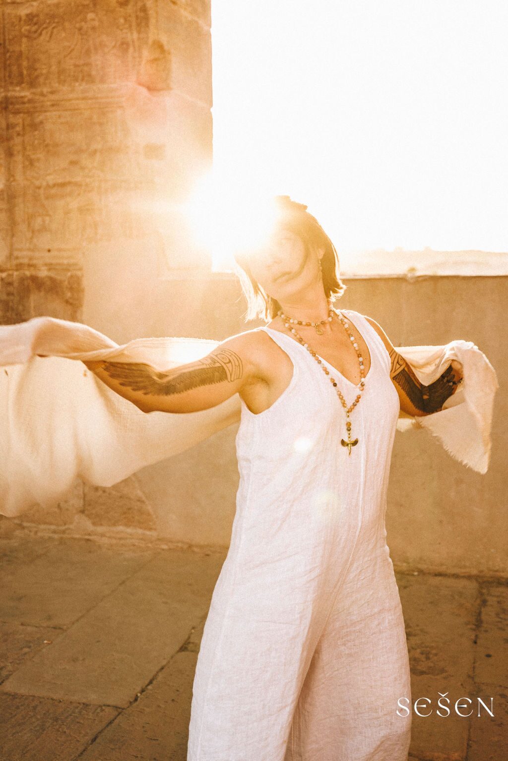 TSA Revna at an ancient Egyptian stone temple, arms outstretched with sheer white fabric billowing like wings, face lifted toward a blazing golden sun flare through the columns